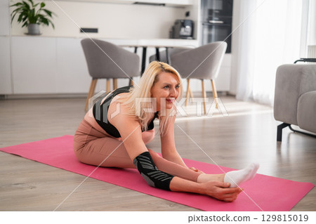 Relaxed blonde middle aged woman sitting on floor stretching to one leg during yoga home practice in stylish apartment 129815019