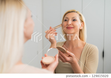 Beautiful female in light clothing using makeup brush to apply facial powder for smooth skin Beautiful female in light clothing using makeup brush to apply facial powder for smooth skin 129815020