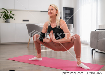 Relaxed blonde woman in middle age doing wide leg squat on yoga mat at home to improve strength, balance, and core stability 129815022