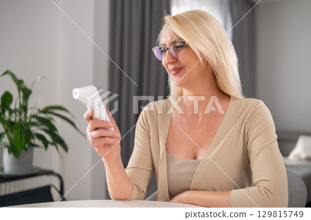Cheerful blonde middle-aged woman in beige outfit looking happily at thermometer while feeling better in modern apartment living room Cheerful blonde middle-aged woman in beige outfit looking happily at thermometer while feeling better in modern apartment living room 129815749