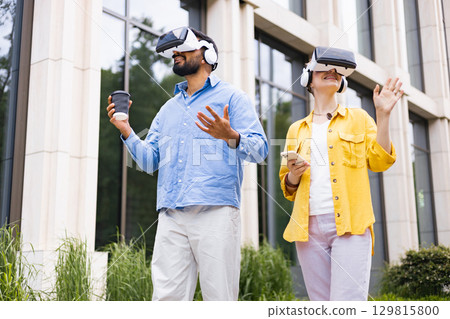 Two people in virtual reality headsets explore the outdoors near a modern building, one with a coffee, the other with a smartphone. 129815800