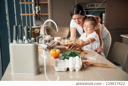 Reads the recipe. Mother with her daughter are preparing food on the kitchen Reads the recipe. Mother with her daughter are preparing food on the kitchen 129817422