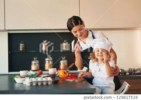 Eating pepper. Mother with her daughter are preparing food on the kitchen Eating pepper. Mother with her daughter are preparing food on the kitchen 129817519
