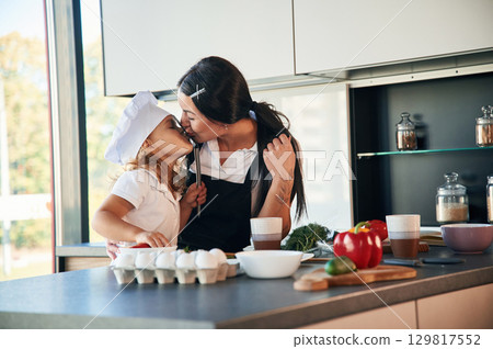 Giving a kiss. Mother with her daughter are preparing food on the kitchen Giving a kiss. Mother with her daughter are preparing food on the kitchen 129817552