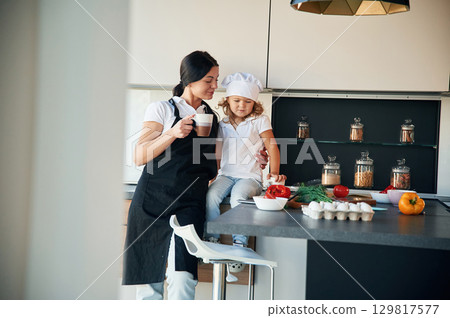 Woman is holding a cup with drink. Mother with her daughter are preparing food on the kitchen 129817577
