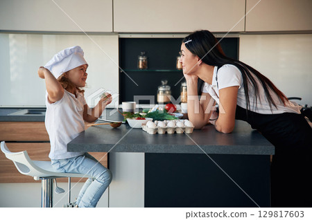 Girl is in the chief hat. Mother with her daughter are preparing food on the kitchen 129817603