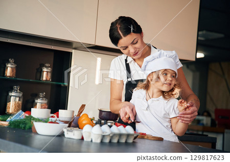 Woman is in apron, girl in chief hat. Mother with her daughter are preparing food on the kitchen Woman is in apron, girl in chief hat. Mother with her daughter are preparing food on the kitchen 129817623