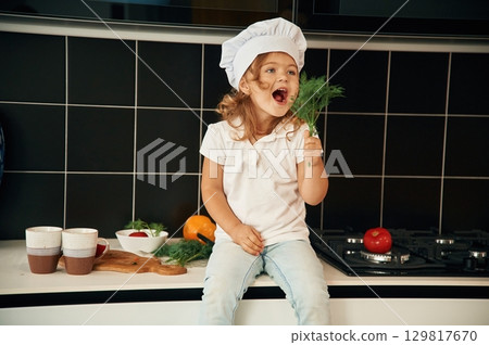 Cheerful little girl in chief hat is sitting on the table of kitchen with vegetables Cheerful little girl in chief hat is sitting on the table of kitchen with vegetables 129817670