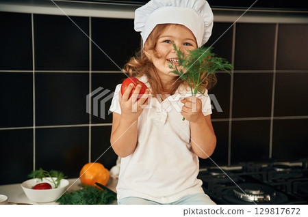 Cheerful little girl in chief hat is sitting on the table of kitchen with vegetables 129817672