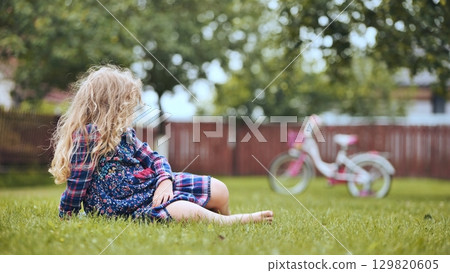 Blond child sitting on verdant lawn, gazing at bicycle while enjoying carefree family moment in sunlit backyard Blond child sitting on verdant lawn, gazing at bicycle while enjoying carefree family moment in sunlit backyard 129820605
