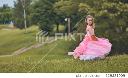 Young girl twirling happily in sunlit green park, wearing pink dress, surrounded by vibrant foliage 129821009