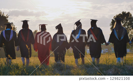 Graduates in robes and caps walking in a field at sunset, holding hands and celebrating their academic success and camaraderie Graduates in robes and caps walking in a field at sunset, holding hands and celebrating their academic success and camaraderie 129821108