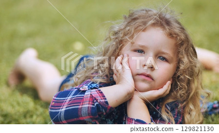 Blonde haired girl relaxing on grassy park lawn, chin propped on hands, radiating childhood innocence and summer delight 129821130