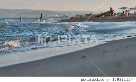 Gentle waves washing Crimean beach at sunset with tourists enjoying summer vacation Gentle waves washing Crimean beach at sunset with tourists enjoying summer vacation 129821134