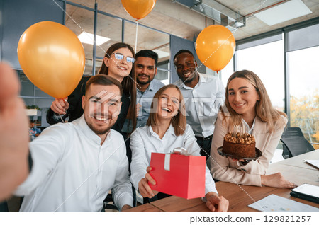Selfie of happy woman with gift. Employee having a birthday in the office, group of workers 129821257
