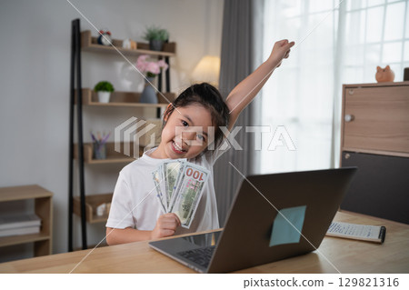Young girl celebrating success while holding cash in a bright home office, showcasing joy and achievement in financial literacy and personal growth. Young girl celebrating success while holding cash in a bright home office, showcasing joy and achievement in financial literacy and personal growth. 129821316