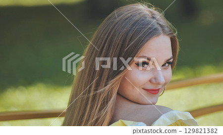 Beautiful young woman with long brown hair, wearing a yellow top, turning and smiling over her shoulder in a park Beautiful young woman with long brown hair, wearing a yellow top, turning and smiling over her shoulder in a park 129821382