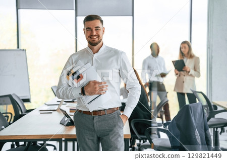 Cheerful man is with documents in hands, standing. Group of office workers are together indoors 129821449