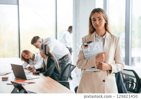 Young woman is holding documents in hands, standing. Group of office workers are together indoors Young woman is holding documents in hands, standing. Group of office workers are together indoors 129821450