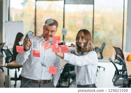 Two people are putting stickers on the glass. Planning the project. Group of office workers are together indoors Two people are putting stickers on the glass. Planning the project. Group of office workers are together indoors 129821508