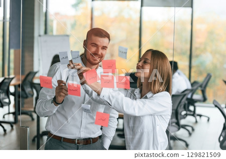 Two people are putting stickers on the glass. Planning the project. Group of office workers are together indoors Two people are putting stickers on the glass. Planning the project. Group of office workers are together indoors 129821509
