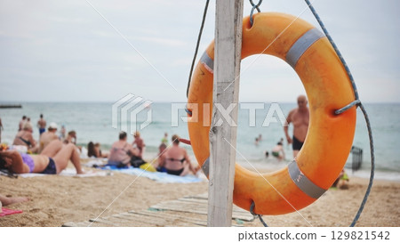 Orange lifebuoy resting on sandy crimean beach, sunbathing individuals surrounding lifesaving ring near black sea shoreline 129821542