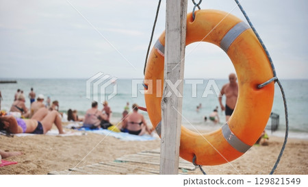 Orange lifebuoy resting on sandy crimean beach, sunbathing individuals surrounding lifesaving ring near black sea shoreline 129821549