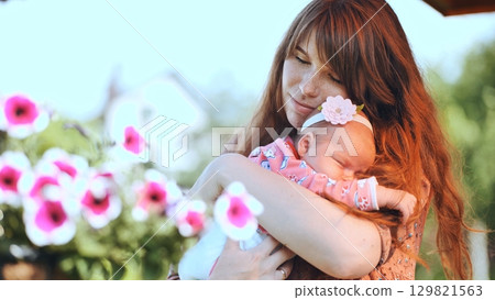 Redheaded mom tenderly embracing sleeping newborn daughter in pink dress, nestled among blooming petunias, sharing intimate family moment Redheaded mom tenderly embracing sleeping newborn daughter in pink dress, nestled among blooming petunias, sharing intimate family moment 129821563