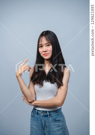 Young woman in casual attire giving a playful point gesture against a light gray background showcasing confidence and personality in a studio setting 129821895