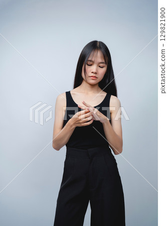 Young woman in sleeveless black top and black pants posing thoughtfully against a simple gray background, showing a contemplative moment in a minimalist setting 129821900