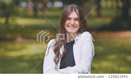 Smiling georgian woman with flowing brown hair, wearing layered white and black tops, standing in sunlit park environment 129821941