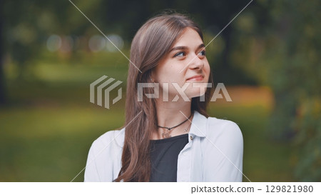 Portrait of a cheerful young woman with long brown hair, wearing a white shirt, smiling and looking up in a park Portrait of a cheerful young woman with long brown hair, wearing a white shirt, smiling and looking up in a park 129821980