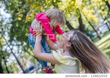 Happy mother lifting her daughter in a sunlit park, sharing joyful moments and laughter on a beautiful day filled with love and fun 129822010