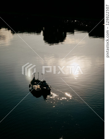 Wooden fishing vessel gliding across tranquil river waters, reflecting golden sunset hues, surrounding landscape mirroring silky water surface with shadowy tree silhouettes. Small fishing boat sailing Wooden fishing vessel gliding across tranquil river waters, reflecting golden sunset hues, surrounding landscape mirroring silky water surface with shadowy tree silhouettes. Small fishing boat sailing 129822507