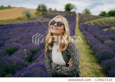 Happy young blonde woman with sunglasses and crossed arms is standing in lavender field, enjoying scent and beautiful purple landscape. Laughing caucasian woman standing amid vibrant purple flowers 129822520