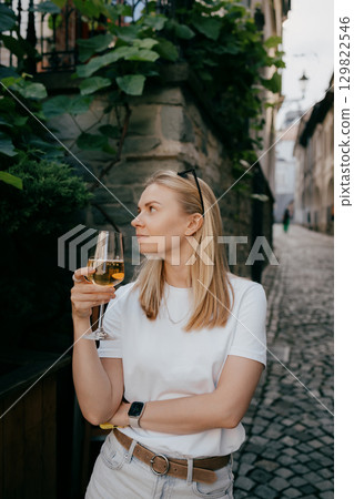 Blonde woman holding glass of white wine while standing on narrow cobblestone street in old European town, enjoying summer vacation and exploring local culture. Blonde woman sipping white wine 129822546