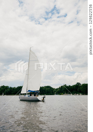 Sailboat gliding across lake, its white sail billowing against backdrop of soft, gray clouds, creating serene and picturesque landscape perfect for relaxation and exploration. Sleek sailboat cruising 129822556