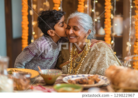 Indian boy kissing his grandmother on the cheek at the festive table in honor of Diwali festival. Traditional decoration of the house with flower garlands and candles. Indian boy kissing his grandmother on the cheek at the festive table in honor of Diwali festival. Traditional decoration of the house with flower garlands and candles. 129822967
