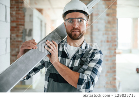 Holding metal plank. Construction worker in uniform in empty unfinished room 129823713