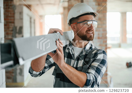 With metal plank in hands. Construction worker in uniform in empty unfinished room 129823720