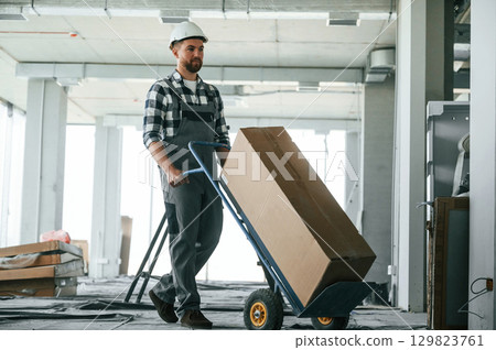 Transporting long paper box. Construction worker in uniform in empty unfinished room Transporting long paper box. Construction worker in uniform in empty unfinished room 129823761