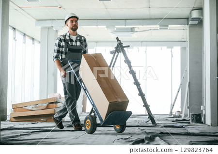 With large box, transporting the material. Construction worker in uniform in empty unfinished room 129823764