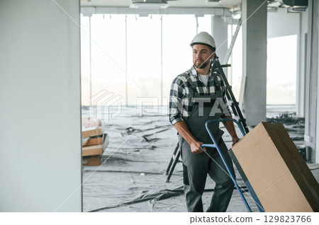 With large box, transporting the material. Construction worker in uniform in empty unfinished room 129823766