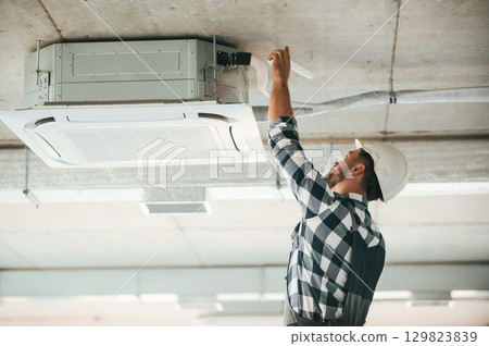 Ventilation system high up on the ceiling, fixing. Repairman in uniform is in the unfinished building 129823839
