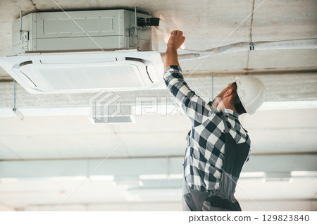 Ventilation system high up on the ceiling, fixing. Repairman in uniform is in the unfinished building 129823840
