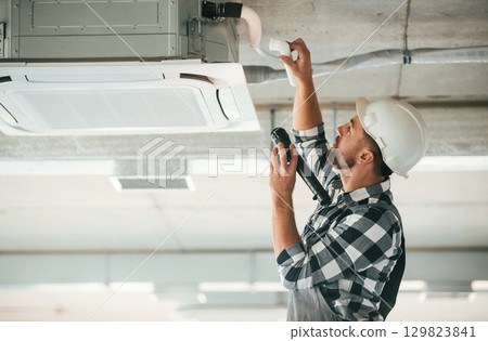 Ventilation system high up on the ceiling, fixing. Repairman in uniform is in the unfinished building Ventilation system high up on the ceiling, fixing. Repairman in uniform is in the unfinished building 129823841