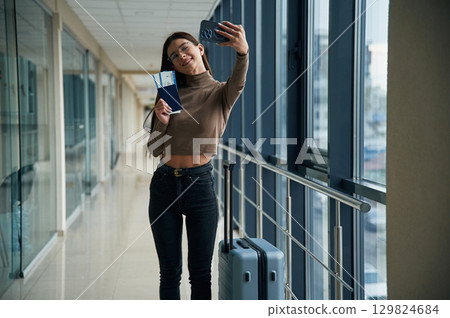 Making selfie with phone, smiling. Young woman traveler in the airport entrance hall Making selfie with phone, smiling. Young woman traveler in the airport entrance hall 129824684