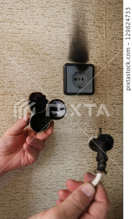 male hands holding scorched wire and tee against background of wall home socket with black traces of burning, dangerous domestic incident related to risk of fire due to malfunction of electrical male hands holding scorched wire and tee against background of wall home socket with black traces of burning, dangerous domestic incident related to risk of fire due to malfunction of electrical 129824733