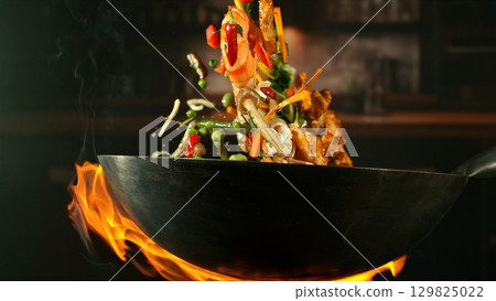 Freeze Motion of Wok Pan with Flying Ingredients in the Air and Fire Flames. Kitchen Interior on Background. 129825022