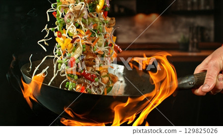 Freeze Motion of Wok Pan with Flying Ingredients in the Air and Fire Flames. Kitchen Interior on Background. 129825026